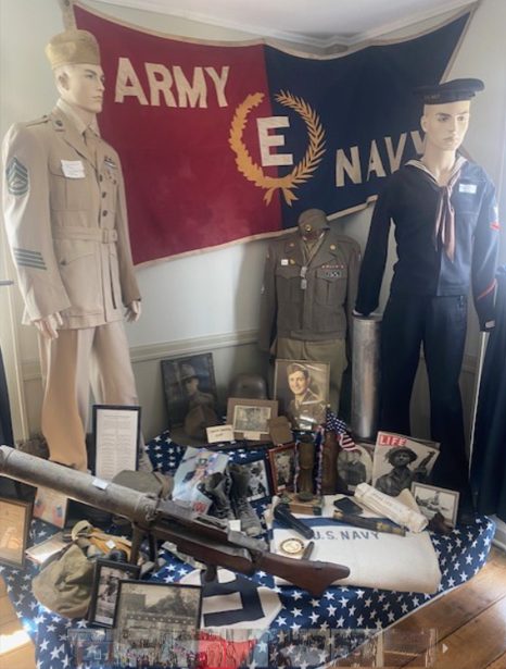 Army and Navy uniforms, a rifle, and wartime keepsakes beneath an Army–Navy E banner